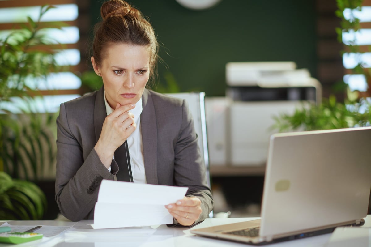Concerned modern middle aged woman employee in modern green office in grey business suit with laptop, showing frustration with job search challenges